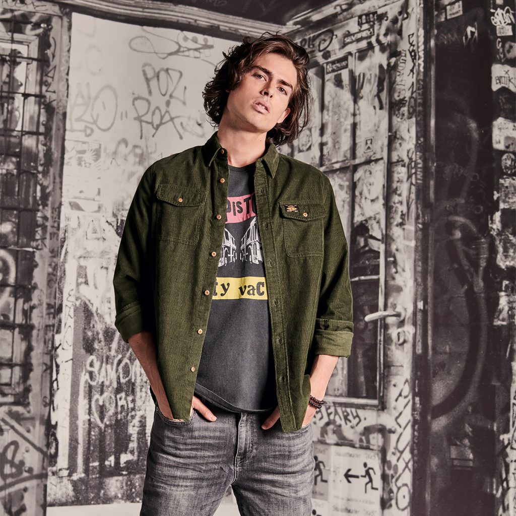 A man in casual Superdry clothes stands in front of graffiti.