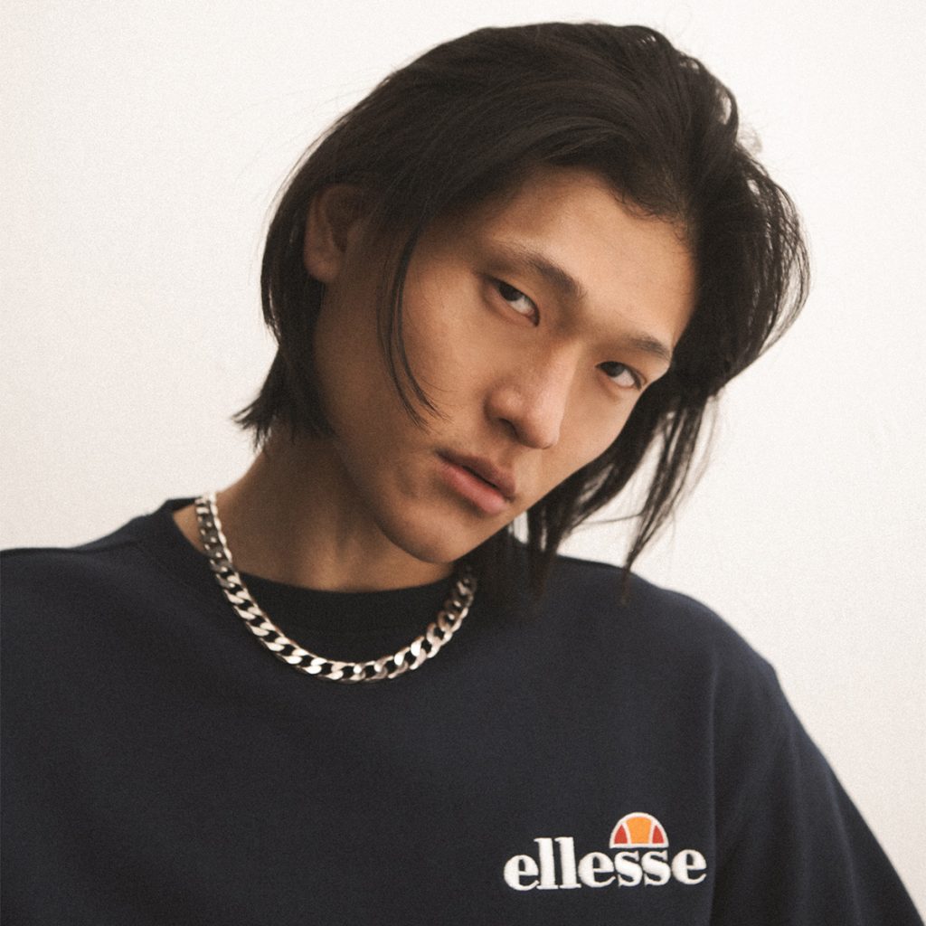 A man wearing an Ellesse sweatshirt and a gold chain looks at the camera.