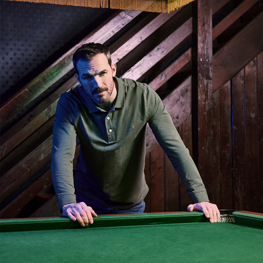 A man leaning on a pool table wearing a green Farah polo shirt