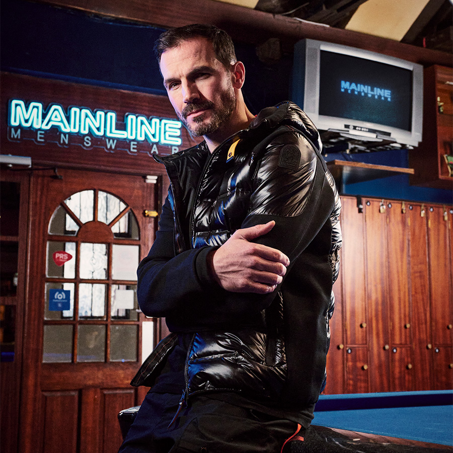 A man leaning on a pool table in front of a Mainline Menswear neon sign